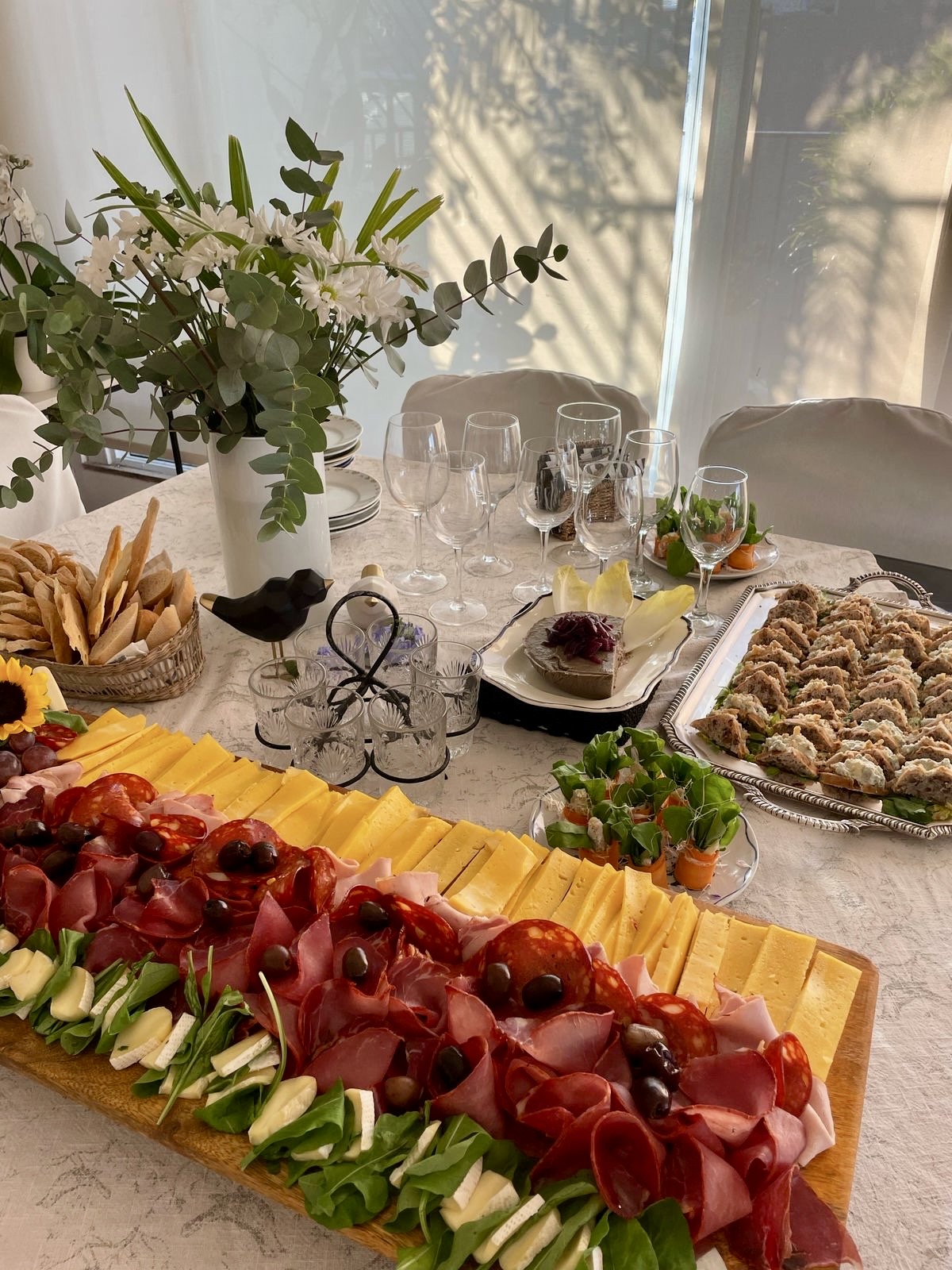 Mesa de catering casera con flores, vajilla, fiambres, quesos y bocados